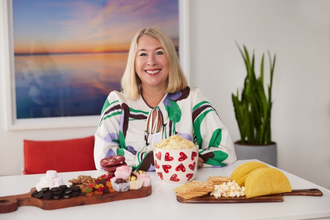 Yume founder Katy Barfield sitting at a table, with an abstract painting behind her and food on table in front of her.