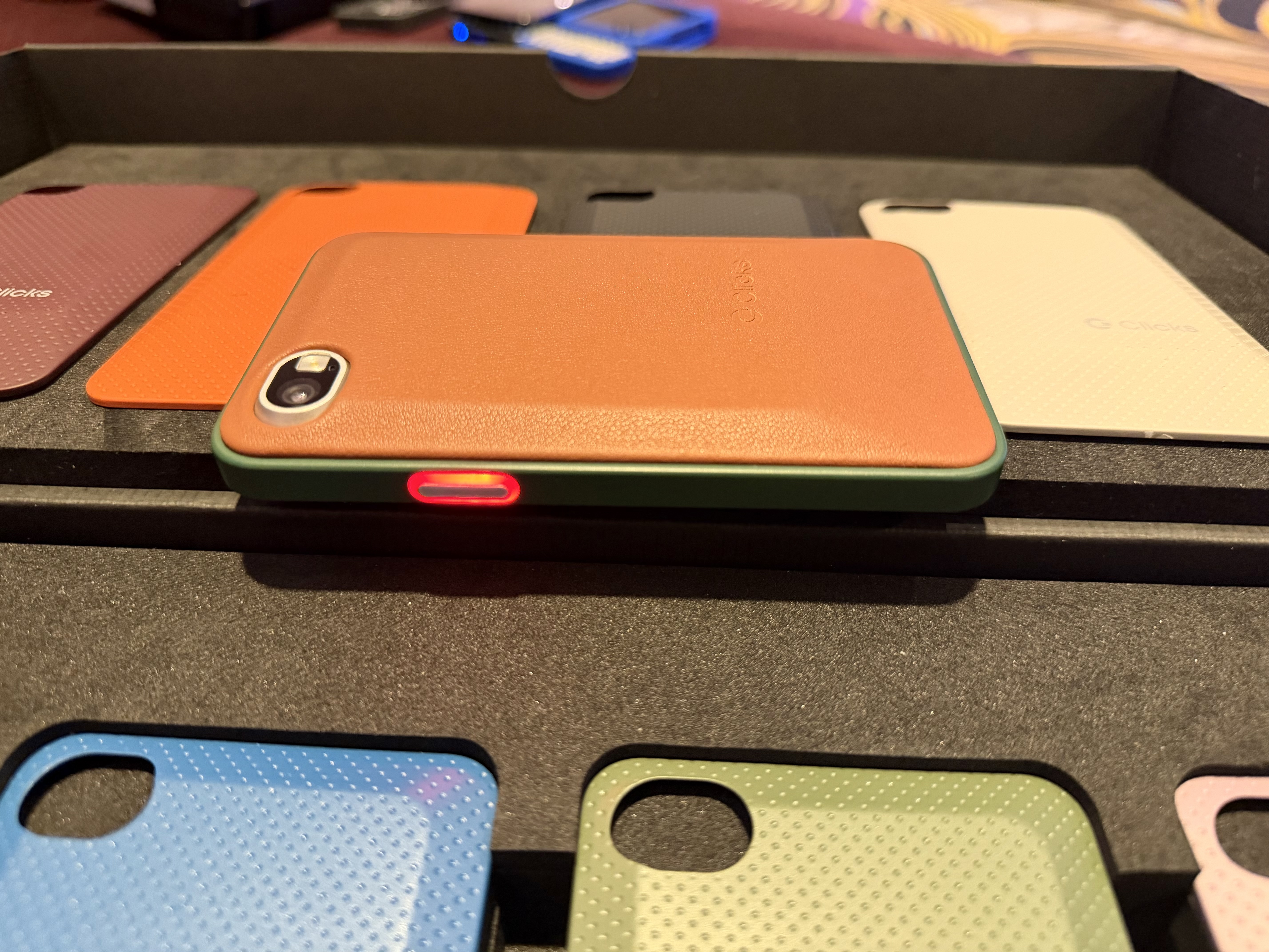 a photo showing the Clicks Communicator device, with an orange shell and a red lit-up notification button on the side.